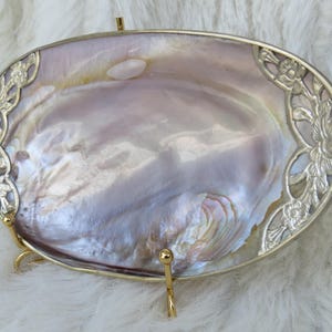 May include: An oval-shaped decorative tray featuring a polished, iridescent mother-of-pearl centre. The tray is framed by a gold-toned metal border with intricate floral cutouts on the left and right sides, supported by a gold-toned metal stand.