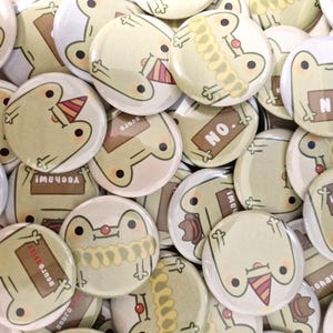 Könnte beinhalten: Eine Sammlung runder Knöpfe mit verschiedenen Designs von Fröschen mit unterschiedlichen Ausdrücken und Texten. Einige Knöpfe haben den Text "NO." darauf, während andere Sätze wie "Yeehaw!" und "Neuro" tragen.