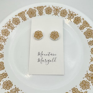 Pode incluir: Um par de brincos de botão de flores brancas e douradas em um cartão branco com o texto "Mountain Marigold". Os brincos são exibidos em um prato branco com um padrão floral dourado na borda.