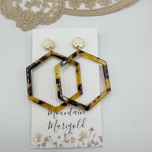 Op de afbeelding: Een paar geometrische zeshoekige oorbellen met een schildpadpatroon. De oorbellen hebben goudkleurige ronde studs. De oorbellen worden getoond op een witte kaart met de tekst "Mountain Marigold".