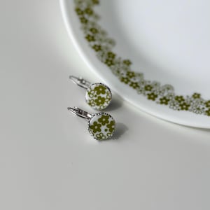 Peut inclure: Une paire de boucles d'oreilles à levier avec un fond blanc et un motif floral dans les tons de vert. Les boucles d'oreilles sont rondes avec une bordure en métal argenté et des fermoirs à levier. Les boucles d'oreilles sont posées sur une surface blanche.