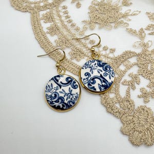 May include: Gold-coloured drop earrings with a circular, porcelain-style design. The earrings have a white background with blue floral patterns and a gold-coloured border. They are displayed on a white surface with a beige lace trim.