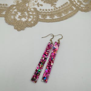 Peut inclure: Une paire de boucles d'oreilles pendantes de forme rectangulaire. Les boucles d'oreilles sont remplies de paillettes roses et d'autres éclats colorés. Les crochets des boucles d'oreilles sont de couleur dorée.