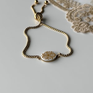 Può includere: Bracciale regolabile color oro con una delicata catena e un motivo floreale centrale bianco e oro. Il bracciale è dotato di una perla scorrevole per la regolazione della taglia. Sfondo neutro.