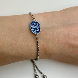 Op de afbeelding: Zilverkleurige armband met een ovale blauw-witte keramische bedel met een bloemmotief. De bedel is gezet in een zilveren rand. De armband heeft een fijne ketting en een verstelbare trekkoordsluiting. De bedel meet ongeveer 2 cm.