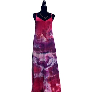 Peut inclure: Longue robe tie-dye dans les tons de rose et de violet, présentée sur un mannequin noir. La robe présente un décolleté en V, des bretelles spaghetti et une coupe longue et fluide. Un collier doré est visible.