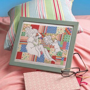 May include: Cross-stitch artwork in a green frame depicts a woman reading with a cat. The background is a patchwork design of various colors. A pair of glasses and a patterned notebook rest nearby. A striped pillow and pink blanket are in the background.
