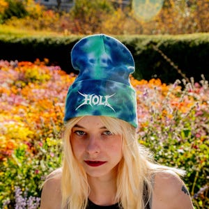 May include: A tie-dye beanie in shades of blue and green, featuring the white word "HOLT" in a stylized font. The hat is made of a ribbed fabric. The background is a colorful garden.