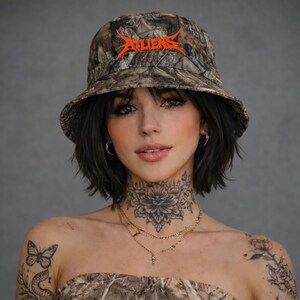 May include: A camouflage bucket hat with the word "ALIENS" in orange lettering. The hat is worn by a person with dark hair and tattoos. The hat is a fashion accessory.