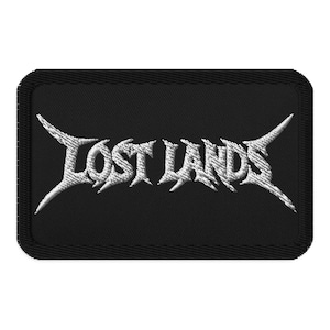 May include: A black rectangular patch with the white embroidered text "LOST LANDS" in a stylized, gothic font. The patch has a black border and is likely designed to be sewn onto clothing or accessories.