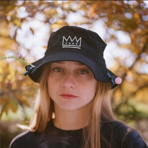 May include: Black bucket hat with a white crown design. The hat has a wide brim and is decorated with a sprig of flowers and a pink ball. The background is a blurred view of autumn foliage.