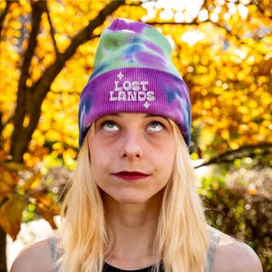 May include: A tie-dye beanie in shades of purple, green, and blue. The beanie features the embroidered text "LOST LANDS" in white. The person in the image is looking up, wearing the beanie.