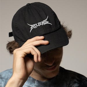 May include: Black baseball cap with the word "ATHENS" embroidered in white, stylized lettering. The cap has a curved brim and an adjustable strap at the back. The cap is made of a durable fabric.