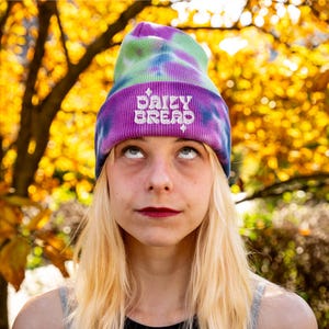 May include: A tie-dye beanie in shades of purple, green, and blue. The hat features the embroidered text "DAILY BREAD" in white. The beanie is worn by a person with blonde hair, against a backdrop of fall foliage.