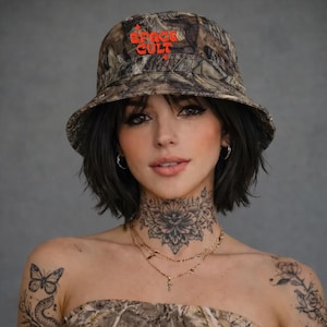 May include: A camouflage bucket hat with the text "SPACE CULT" in red lettering. The hat is worn by a person with dark hair and tattoos on their neck and arm. The person is wearing a matching camouflage top.