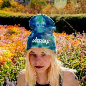 May include: A tie-dye beanie in shades of blue and green, with the word "MERSIU" in white script across the front. The hat is made of a soft, ribbed fabric. The background features colorful flowers and foliage.