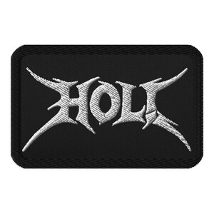 May include: Black embroidered patch with the white word "HOLY" in a stylized, gothic font. The patch has a rectangular shape with rounded corners. The letters have sharp, pointed serifs, giving it a heavy metal aesthetic.