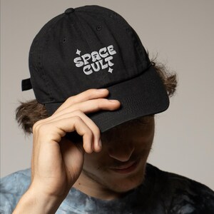 May include: Black baseball cap with the text "SPACE CULT" in a white, stylized font. The cap has a curved brim and a small adjustable strap at the back. The cap is made of a soft, durable material.