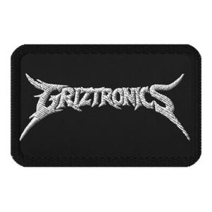 May include: A rectangular black embroidered patch featuring the word GRIZTRONICS stitched in white thread. The lettering is designed in a sharp, angular, metallic-style font with pointed extensions on the first and last letters of the text.