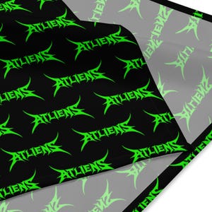 May include: Black fabric with a repeating neon green "ATLIENS" text pattern. The text has a stylized, sharp font. The fabric is folded, revealing a white underside.