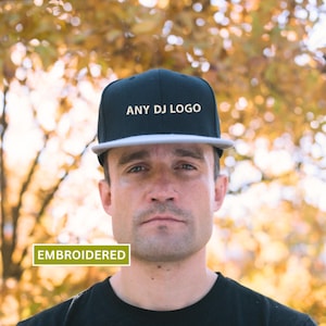 May include: Black and gray flat-brimmed cap with the text "ANY DJ LOGO" embroidered in white. The cap has a gray brim and the word "EMBROIDERED" in a green rectangle. The background is a blurred view of autumn foliage.