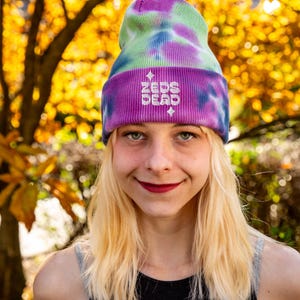 May include: A tie-dye beanie in shades of purple, green, and blue. The hat features the text "ZEDS DEAD" embroidered in white. The person wearing the hat has blonde hair and is smiling. The background is a blurred view of fall foliage.