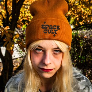 May include: Mustard yellow beanie with the text "SPACE CULT" in black, with a small star design. The beanie is made of a soft, knitted material. The beanie is worn by a person with blonde hair.