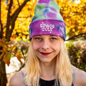 May include: A tie-dye beanie in shades of purple, green, and blue. The beanie features the embroidered text "SPACE CULT" in white. The person in the image is wearing the beanie and a black top.