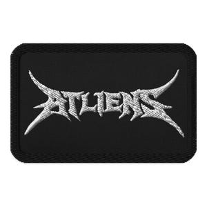 May include: Black rectangular patch with the word "ATLIENS" embroidered in white, stylized, gothic lettering. The patch has a black border and is likely for clothing or accessories.