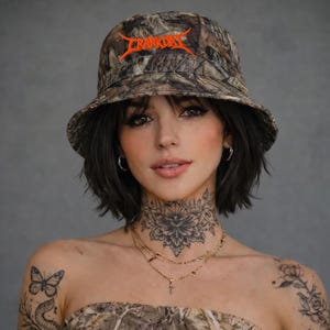 May include: A camouflage bucket hat with the word "CRANKDAY" in orange lettering. The hat is worn by a person with dark hair and tattoos on their neck and shoulder. The hat is made of a camouflage pattern fabric.