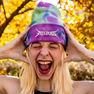 May include: A tie-dye beanie in shades of purple, green, and blue. The beanie features the word "ATHENS" embroidered in silver across the front. The background is a blur of yellow and orange.
