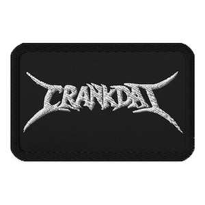 May include: Black rectangular patch with the word "CRANKDAT" embroidered in white, stylized with sharp, angular fonts and extended serifs. The patch has a black border.