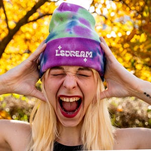 May include: A tie-dye beanie in shades of purple, green, and blue. The beanie features the word "LSDREAM" embroidered in white. The person in the image is wearing the beanie and has their hands on their head.