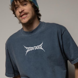 May include: A blue-grey t-shirt with the text "ZEDS DEAD" in a stylized white font. The shirt has a crew neck and short sleeves. The image shows a person wearing the shirt.