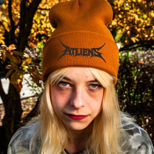 May include: Mustard yellow knit beanie with the word "ALIENS" in a black, stylized font. The beanie is worn by a person with blonde hair and red lipstick, set against a blurred background of fall foliage.