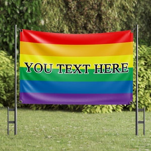 May include: A rainbow flag with the text "YOU TEXT HERE" printed in black on the green stripe. The flag is attached to a black metal pole and is displayed in a grassy area.