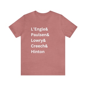 May include: A short-sleeved, crew-neck t-shirt in a dusty rose hue. The shirt features the names L'Engle, Paulsen, Lowry, Creech, and Hinton printed in white, stacked vertically down the front.