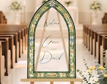 May include: A clear acrylic wedding welcome sign with a stained-glass style border, displaying "Welcome" and the names "Grace and David." The sign is on a wooden easel in a church, with floral arrangements and pews in the background.