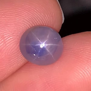 May include: A round, light purple gemstone with a six-pointed star pattern. The gem is held between fingers, showcasing its polished surface and the star effect created by light refraction within the stone. The gem's colour is a soft lavender.