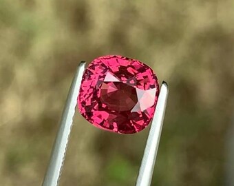 May include: A vibrant pink gemstone with a cushion cut, held by tweezers against a blurred green background.