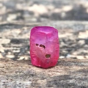 May include: A small, cylindrical, vibrant pink gemstone bead with a faceted surface. The stone features natural inclusions and dark, irregular spots on its exterior, showcasing a translucent quality and a polished, slightly uneven texture throughout.