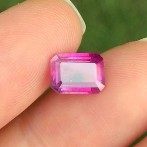 May include: A vibrant pink gemstone with an emerald cut. The gemstone is faceted and has a slight iridescent sheen.