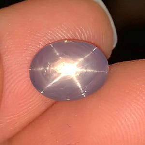 May include: A close-up of a lavender-colored star sapphire gemstone. The oval-shaped stone has a six-rayed star pattern radiating from a central point of light. The gem is held between two fingers.