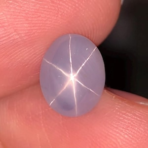 May include: Oval-shaped, lavender-colored gemstone with a six-rayed star pattern. The star effect is created by light reflecting off of inclusions within the stone. The gem is held between fingers, showcasing its polished surface and unique optical phenomenon.
