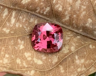 May include: A vibrant pink gemstone with a cushion cut, displayed on a brown leaf with white markings.
