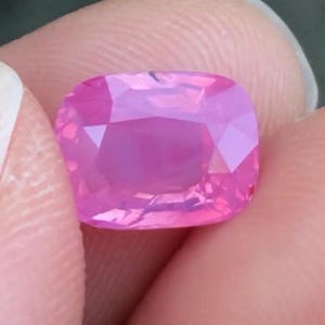 May include: A faceted, cushion-cut pink gemstone. The gem is held between fingers, showcasing its vibrant colour and intricate facets. The stone's pink hue ranges from light to deep, with a polished surface.