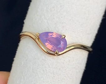 Puede incluir: Un anillo de oro con una piedra preciosa de color morado claro en forma de pera montada en una banda curva. La piedra está sujeta por dos pequeñas garras y la banda de metal tiene un acabado liso y pulido que sigue el contorno de la gema.