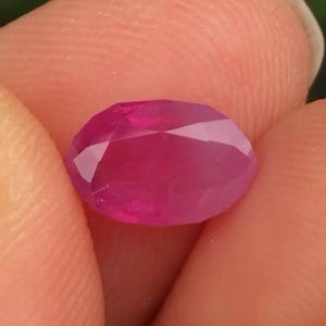 May include: A faceted, oval ruby gemstone. The ruby is a vibrant, deep pinkish-red colour, with multiple facets that reflect light. The gemstone is held between two fingers, showcasing its polished surface and clarity.