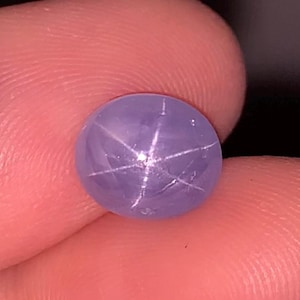 May include: A round, cabochon-cut lavender star sapphire gemstone featuring a distinct six-rayed asterism effect across its smooth, polished surface. The stone displays a translucent light purple hue with a centered white star pattern.