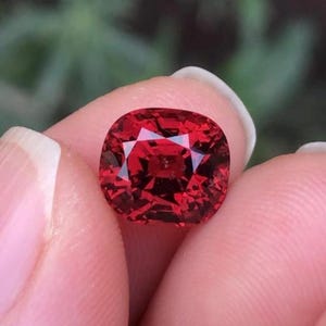May include: A deep red gemstone with a cushion cut. The gemstone is held in a hand.